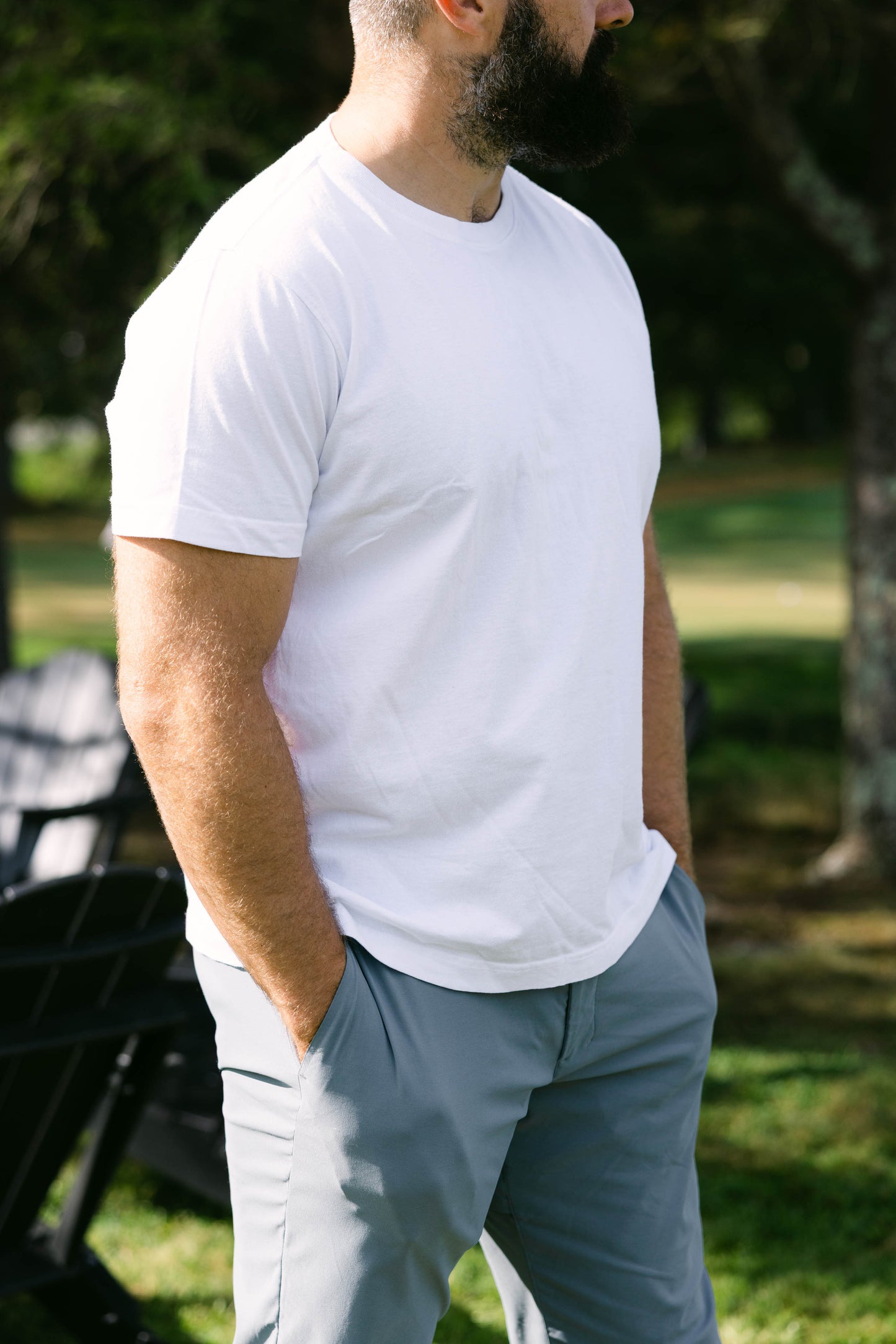 Man wearing a white t-shirt and gray pants standing outdoors with trees and grass in the background
