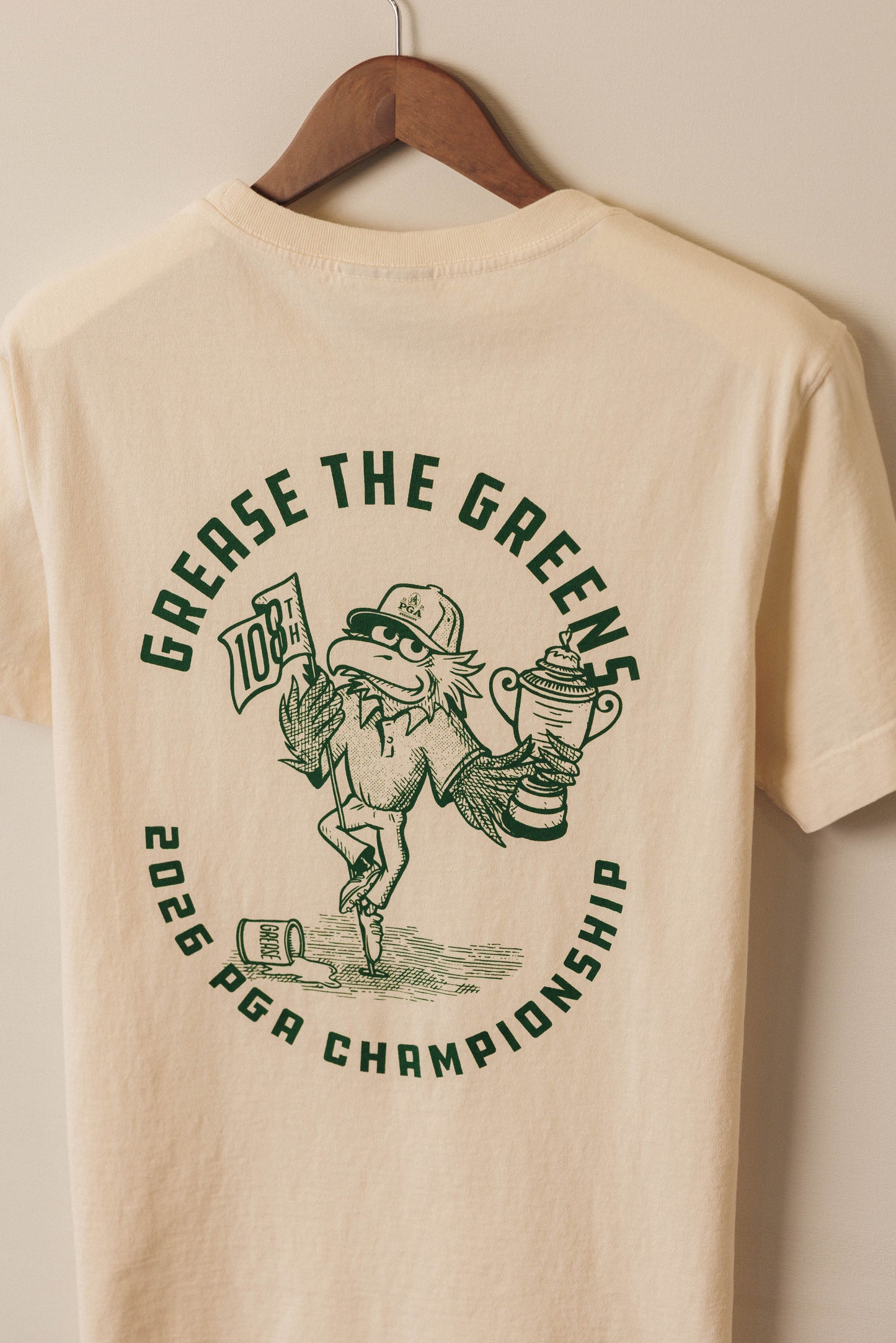 Beige t-shirt with green graphic and text on a hanger against a beige wall