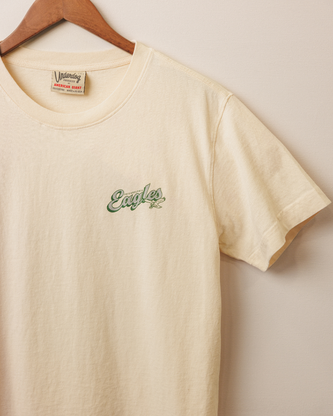 Beige t-shirt with green logo on a hanger against a white background