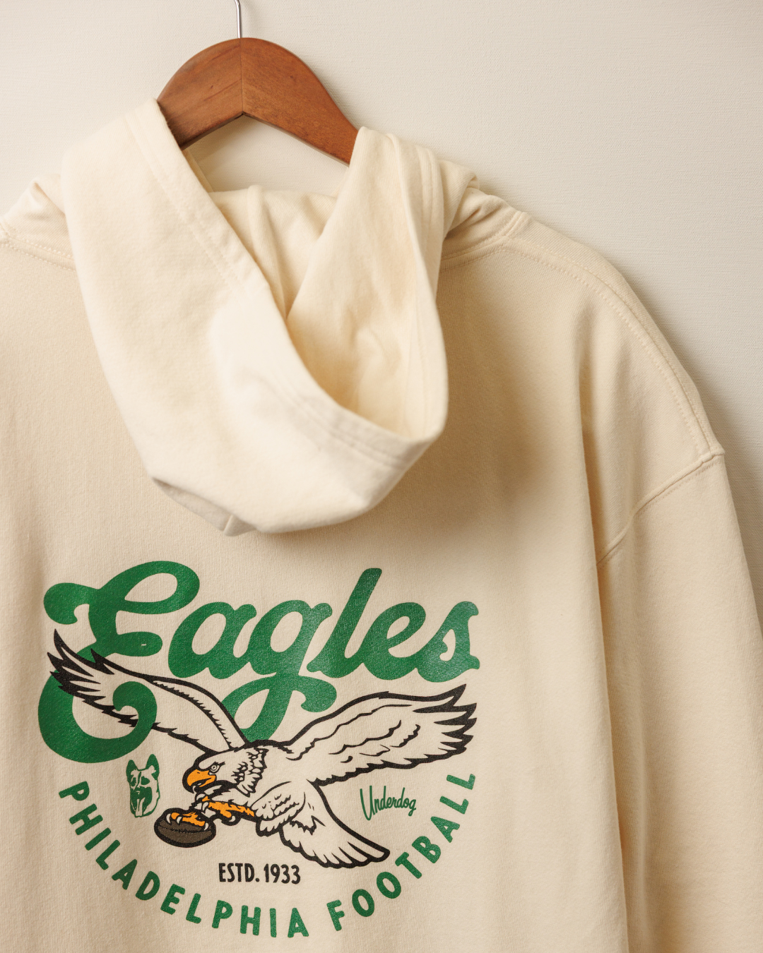 Beige hoodie with green Eagles logo on a wooden hanger against a beige wall