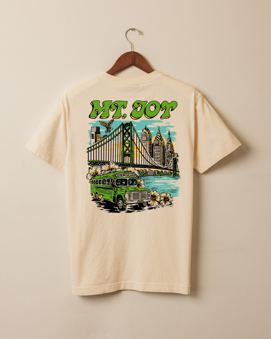 A cream-colored short-sleeve t-shirt hanging on a wooden hanger against a beige wall. The back of the shirt features a large graphic print with the text 'MT. JOY'