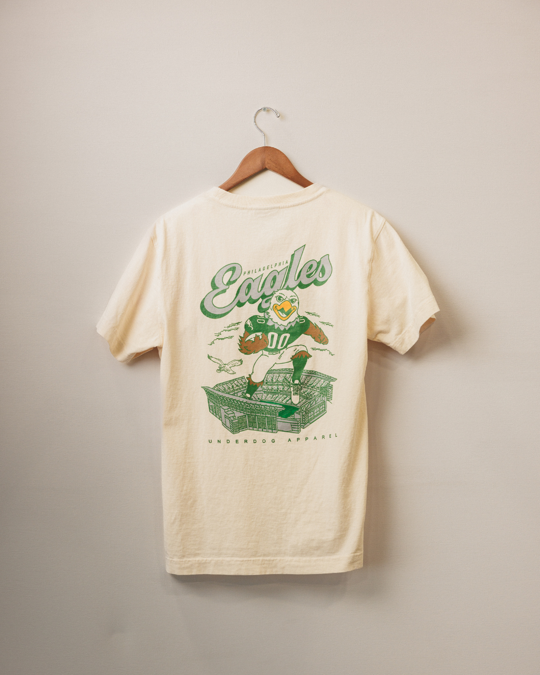 Beige t-shirt with green graphics and text on a hanger against a beige wall