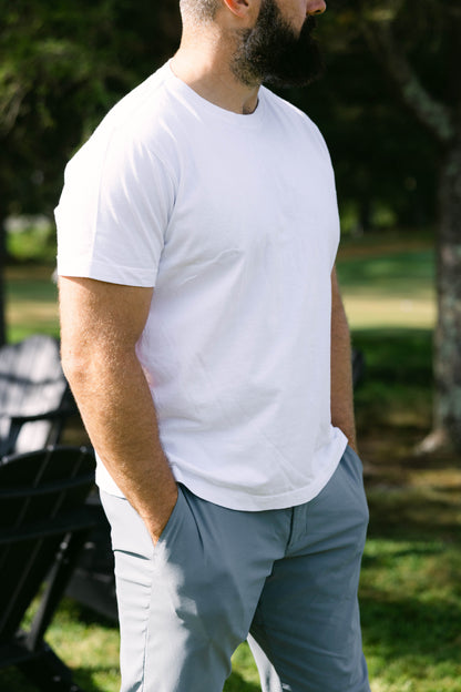 Man wearing a white t-shirt and gray pants standing outdoors with trees and grass in the background
