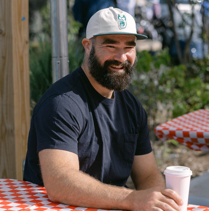 A smiling bearded man wearing a white baseball cap with a green logo and a dark navy t-shirt sits at an outdoor picnic table with a red and white checkered tablecloth, holding a white paper coffee cup.