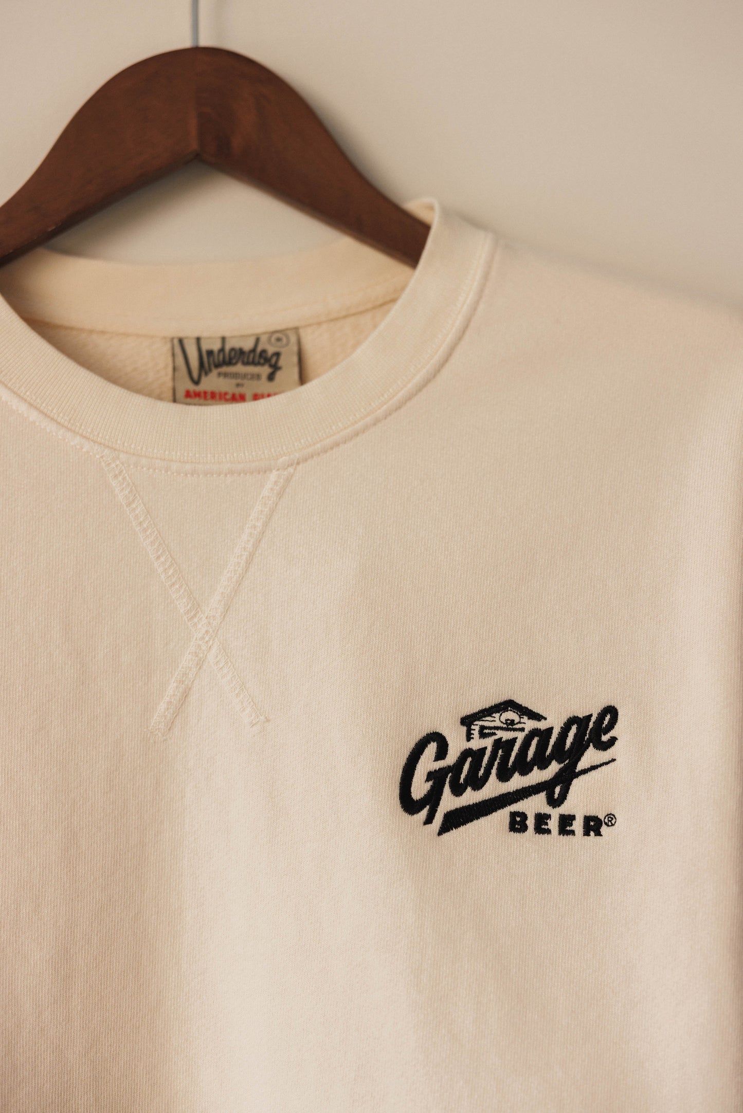 Beige sweatshirt with 'The Garage Beer' logo on a hanger against a white wall.