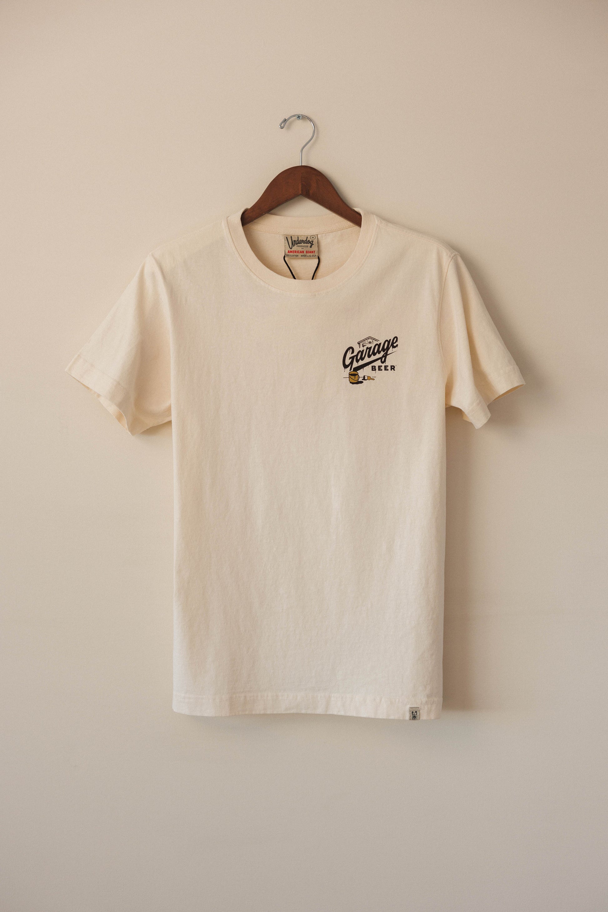 Beige t-shirt with a logo on a wooden hanger against a beige wall