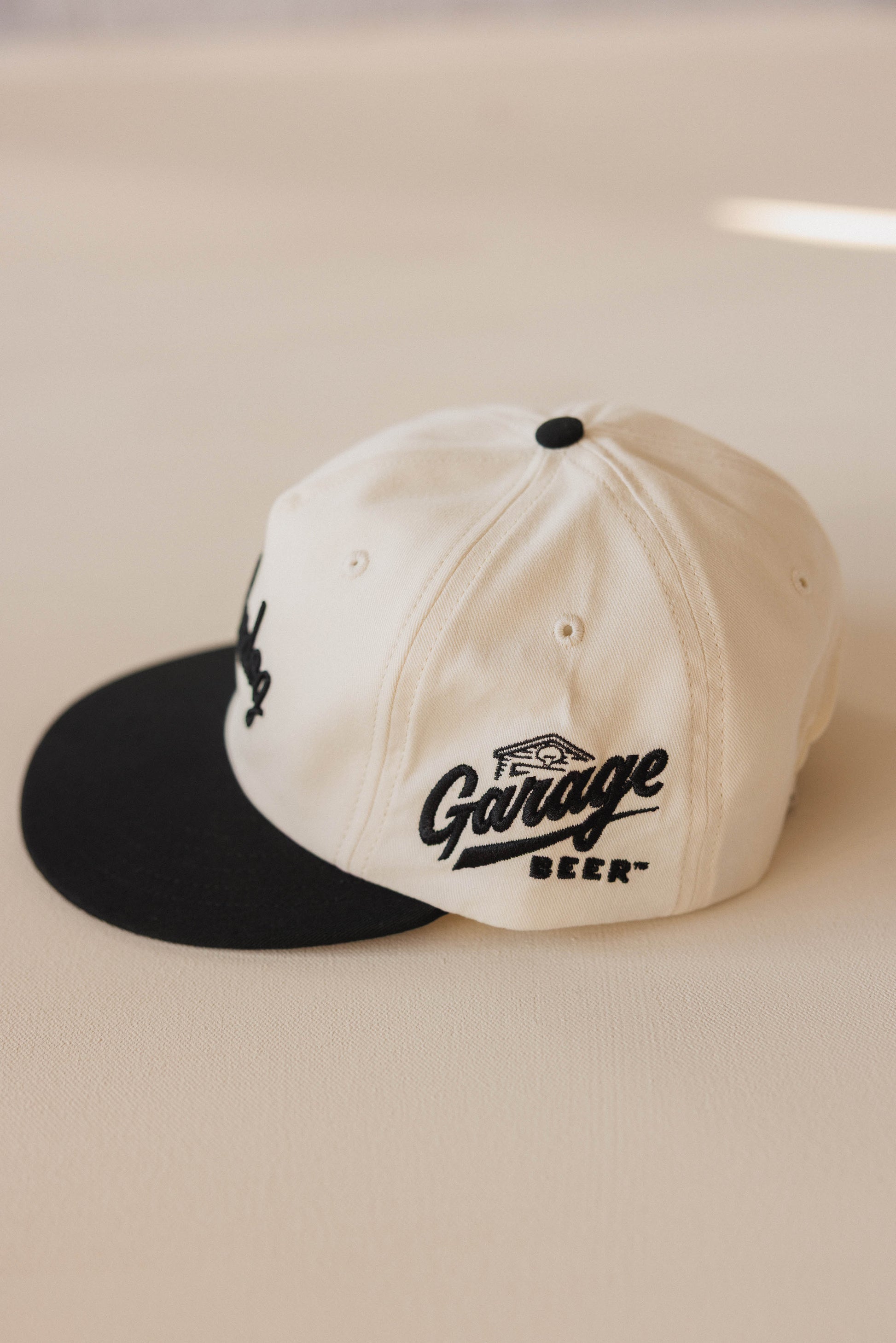 Two-tone cap with black visor and white主体, featuring 'Garage Beer' branding on a beige background.