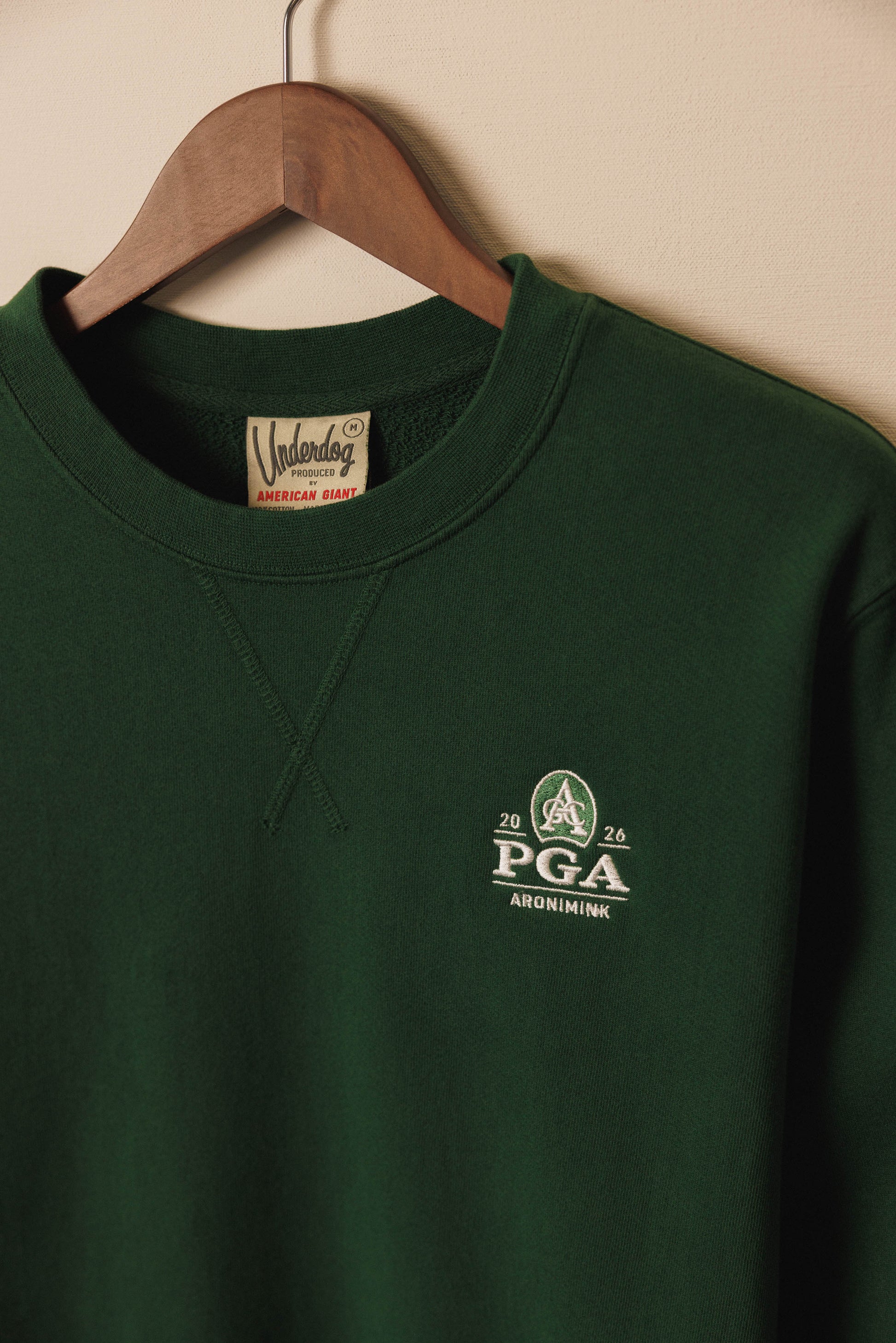 Green sweatshirt with PGA logo on a hanger against a beige wall