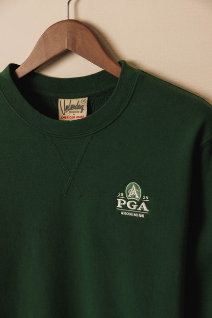 Green sweatshirt with PGA logo on a hanger against a beige wall