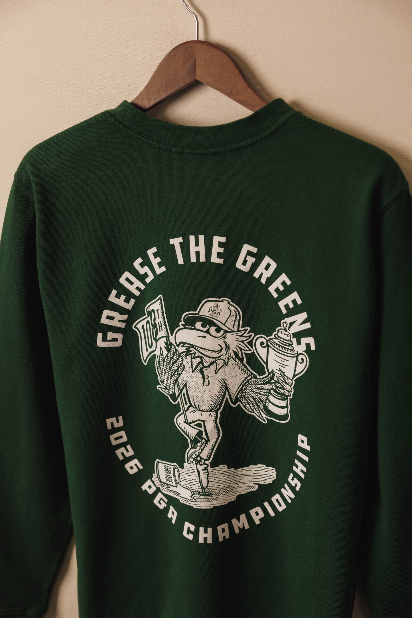 Green sweatshirt with graphic design and text on a hanger against a beige wall