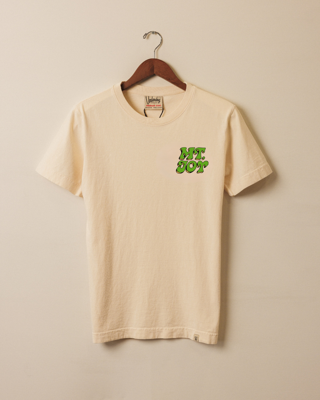 A cream-colored cotton t-shirt hanging on a wooden hanger against a neutral beige wall. The shirt features a green textured graphic on the left chest that reads 'MT JOY'.