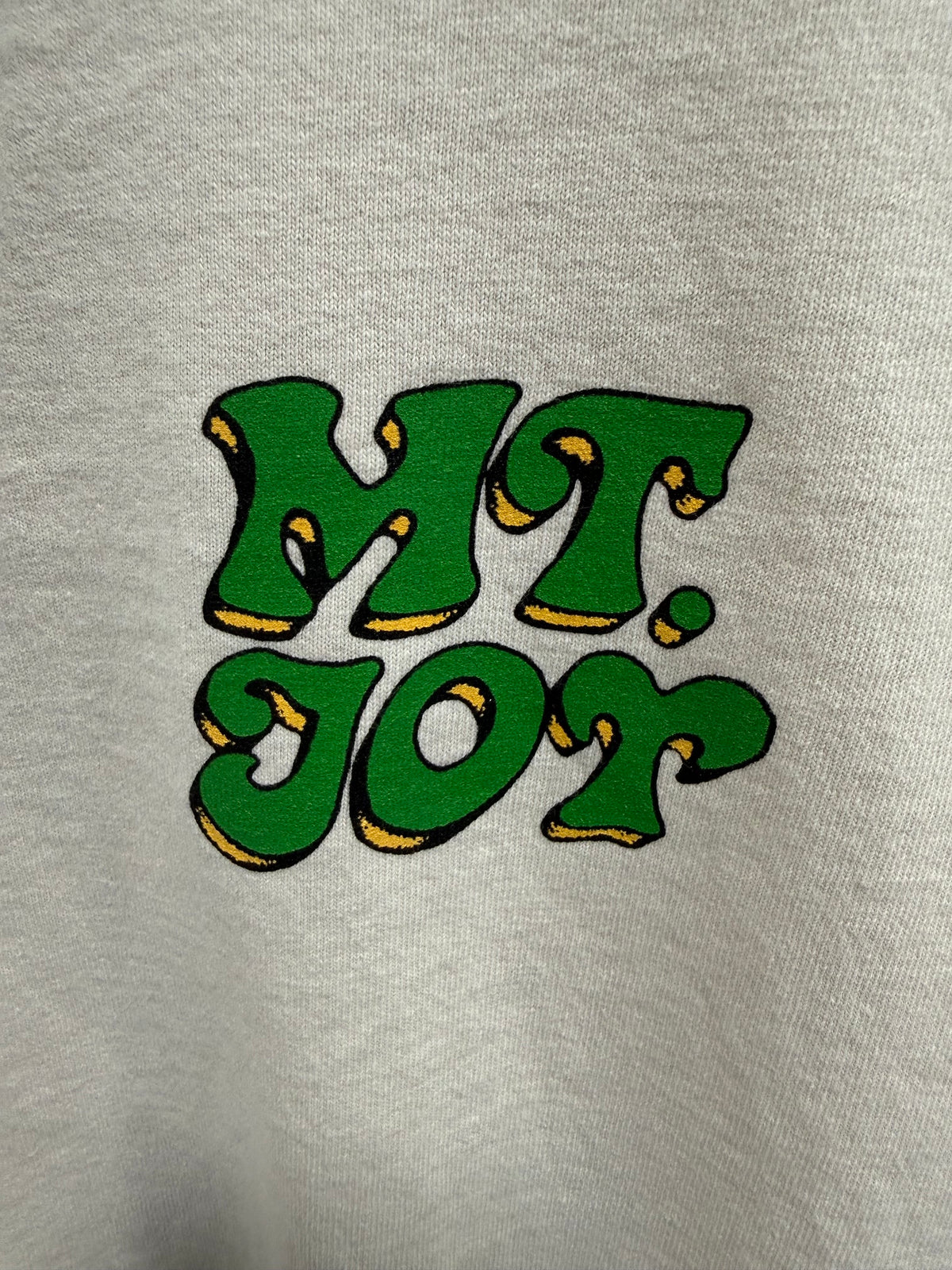 Close-up of a light gray heathered fabric with a large green embroidered graphic reading 'MT. JOT' outlined in black and accented with yellow edges.