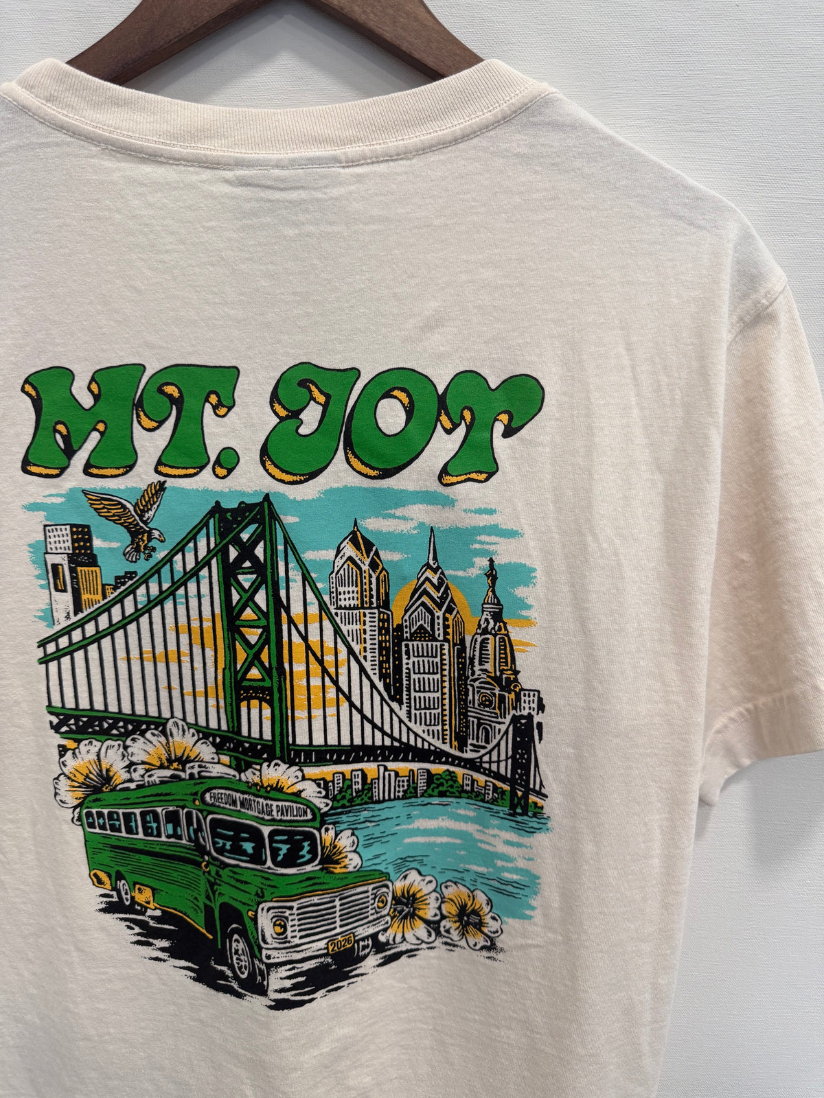An off-white short-sleeve t-shirt hanging on a wooden hanger, featuring a colorful graphic print of the Philadelphia skyline with the text 'MT. JOY' in green, a suspension bridge, a green bus, and yellow flowers.